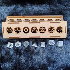 Puede incluir: Una caja de madera con formas geométricas recortadas en la parte delantera y formas geométricas de cristal transparente debajo. La caja tiene una tapa con recortes circulares. Las formas de cristal incluyen una pirámide, un cubo y otros poliedros.