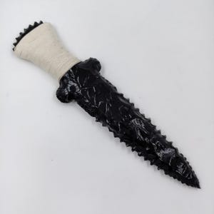 May include: A black obsidian dagger with a sharp, serrated blade and a handle wrapped in off-white cord. The blade features a glossy, reflective surface with distinct knapped edges, while the handle base has a flared, notched design.