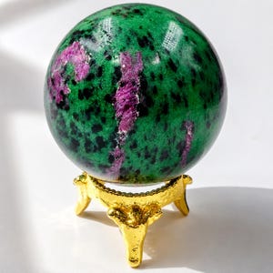 Puede incluir: Una esfera de piedra preciosa pulida, de color verde con motas negras y vetas magenta. Descansa sobre un pequeño soporte dorado ornamentado. Esta esfera es un objeto decorativo, ideal para la decoración del hogar o como regalo único. El soporte mide unos 5 cm de altura.