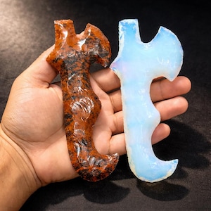 May include: Two carved stone axe pendants. One is a reddish-brown color with black inclusions, and the other is a translucent, opalescent white. The pendants are held in a hand against a dark background.