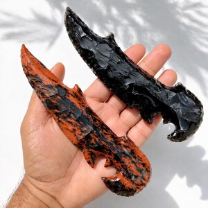 May include: Two obsidian blades with jagged edges. One blade is solid black, the other is a reddish-brown color with black marbling. The blades are held in a hand, showcasing their craftsmanship and unique design. These are likely decorative or ceremonial items.