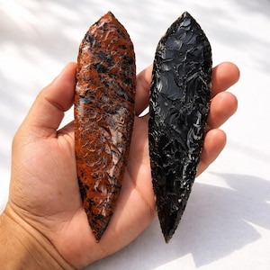 May include: Two hand-held, elongated, obsidian arrowheads. One is a mottled reddish-brown with black inclusions, the other is a solid, glossy black. Both have sharp, pointed tips and are held against a light background.