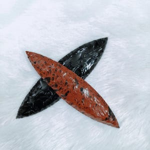 May include: Two obsidian arrowheads. One is a deep, glossy black, and the other is a reddish-brown with black speckles. The arrowheads are crossed against a white, textured background, showcasing their sharp, pointed design and natural stone patterns.