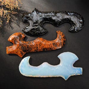 May include: Three hand-carved obsidian and opalite artifacts. The top piece is black, the middle is orange with black marbling, and the bottom is a translucent blue-white. Each has a unique, curved shape with a textured surface.