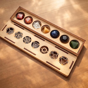 Pode incluir: Um conjunto de caixas de madeira com sete pedras preciosas coloridas, incluindo vermelho, laranja, transparente, amarelo, azul, preto e verde. Abaixo, uma bandeja de madeira com recortes geométricos. O conjunto é exibido em uma superfície de madeira.