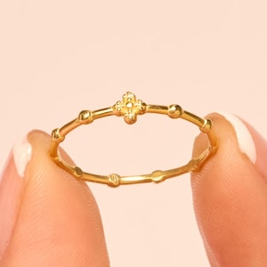 May include: A delicate gold ring featuring a floral design at the center. The ring has a thin band with small, evenly spaced decorative elements. The ring is held between two fingers against a soft pink background.