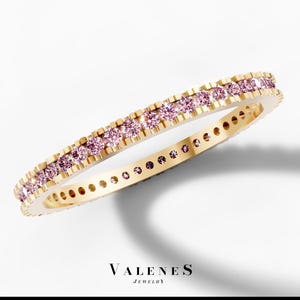 May include: A gold band ring featuring a continuous row of small, round, light pink gemstones set along the top edge. The inner side of the band displays a series of circular cutouts. The text VALENES JEWELRY is printed below the ring.