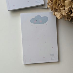 Puede incluir: Bloc de notas gris claro con una ilustración de una nube azul sonriente y corazones rosas. El texto "Keine Angst. Ist nur Liebe." está impreso en la esquina inferior derecha del bloc de notas.