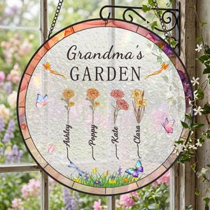 Puede incluir: Letrero de jardín redondo de cristal con el texto "Grandma's GARDEN". El letrero presenta flores coloridas, mariposas y los nombres Ashley, Poppy, Kate y Clara. Cuelga de un soporte de metal negro.
