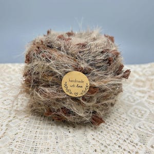 May include: A ball of textured yarn in shades of brown, tan, and silver. The yarn has a fluffy, fuzzy appearance with darker brown accents. A small, round, brown sticker with the words "handmade with love" and a heart is attached to the yarn.