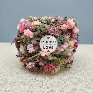 May include: A ball of textured yarn in shades of pink, green, and brown with a circular white sticker on the front that features two red hearts and the text "HAND MADE WITH LOVE" printed in black capital letters in the centre of the yarn ball.