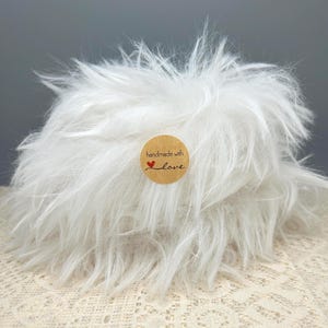 May include: A fluffy white faux fur accessory with a round brown paper sticker centred on the front. The sticker features the black printed text "handmade with" followed by a small red heart icon and the word "love" in cursive script.