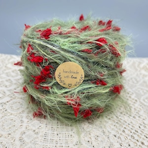 May include: A ball of fuzzy, light green yarn with small, bright red textured accents throughout. A circular, tan-colored paper sticker is attached to the center, featuring the printed text "handmade with love" and a small heart icon.