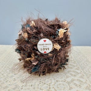 Könnte beinhalten: Ein runder Ball aus dunkelbraunem, flauschigem Garn mit eingestreuten beigen und dunkel-türkisfarbenen Fasern. Ein runder weißer Aufkleber in der Mitte trägt den Text HAND MADE WITH LOVE sowie zwei kleine rote Herzsymbole oben und unten.