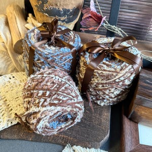 May include: Three round skeins of yarn featuring a blend of brown, blue, and cream fibers with integrated sequins. Each skein is tied with a smooth, dark brown satin ribbon. The yarn has a textured, multi-tonal appearance with visible metallic accents.