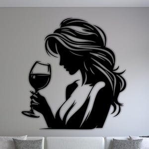Pode incluir: Arte de parede de silhueta preta de uma mulher segurando uma taça de vinho. A obra de arte é montada em uma parede branca acima de um sofá branco e uma mesa de centro preta. O quarto tem um design moderno.