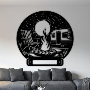 以下が含まれることがあります： 黒い金属製の壁掛けアートで、円の中にキャンプのシーンが描かれています。 デザインには、キャンプファイヤー、折りたたみ椅子、キャンピングカー、満月が含まれています。 アート作品は、灰色のソファの上にある白い壁に取り付けられています。