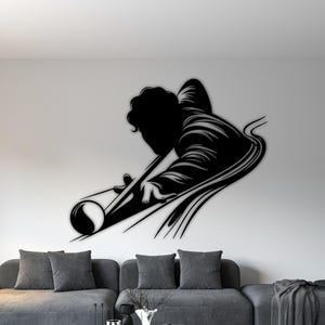Puede incluir: Arte mural de metal negro que representa a una persona jugando al billar. La silueta muestra a un jugador inclinado sobre una mesa de billar, con el taco en la mano, con un diseño fluido. La obra de arte está montada en una pared blanca sobre un sofá gris.