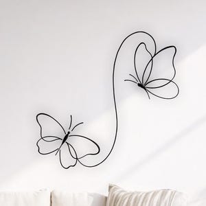 Könnte beinhalten: Schwarze Metall-Wandkunst mit Schmetterlingen. Das Kunstwerk zeigt ein minimalistisches Design von zwei Schmetterlingen, die durch eine geschwungene Linie verbunden sind. Die Schmetterlinge sind an einer weißen Wand über einem Sofa mit neutralfarbenen Kissen befestigt.