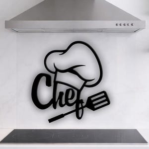 May include: Black metal chef wall art featuring a chef's hat, the word "Chef", and a spatula. The art is mounted on a white wall above a black stovetop in a kitchen setting. Shelves with plants and kitchen utensils are visible.