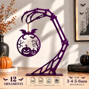 Pode incluir: Um suporte de mão esquelética roxa segurando um ornamento circular com duas abóboras, morcegos e árvores sem folhas. O ornamento está pendurado por um cordão. O design inclui texto indicando 12 ornamentos e material de 3-4-5-6mm.