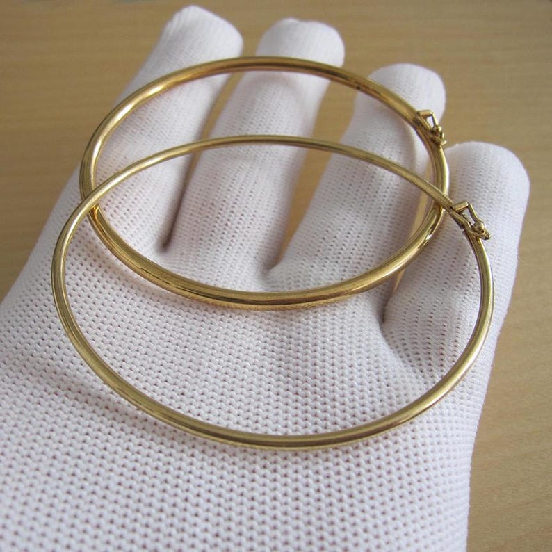 May include: Two gold-colored, thin, round bangles are displayed. The bangles are made of a shiny, metallic material. The bangles are resting on a white, textured surface. The bangles have a simple, elegant design.