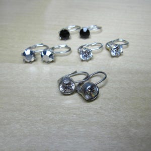 May include: Several pairs of leverback earrings with various gemstone settings are displayed. The earrings feature silver-toned metal and include clear, black, and faceted gemstones. The earrings are arranged on a light-colored wooden surface.