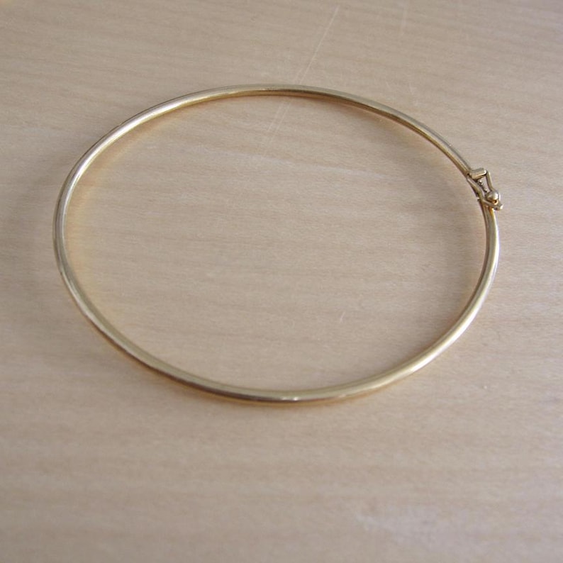 May include: A gold-colored bangle bracelet. The bracelet is a simple, circular design with a clasp. The metal has a shiny finish. The bracelet is shown on a light-colored wooden surface.
