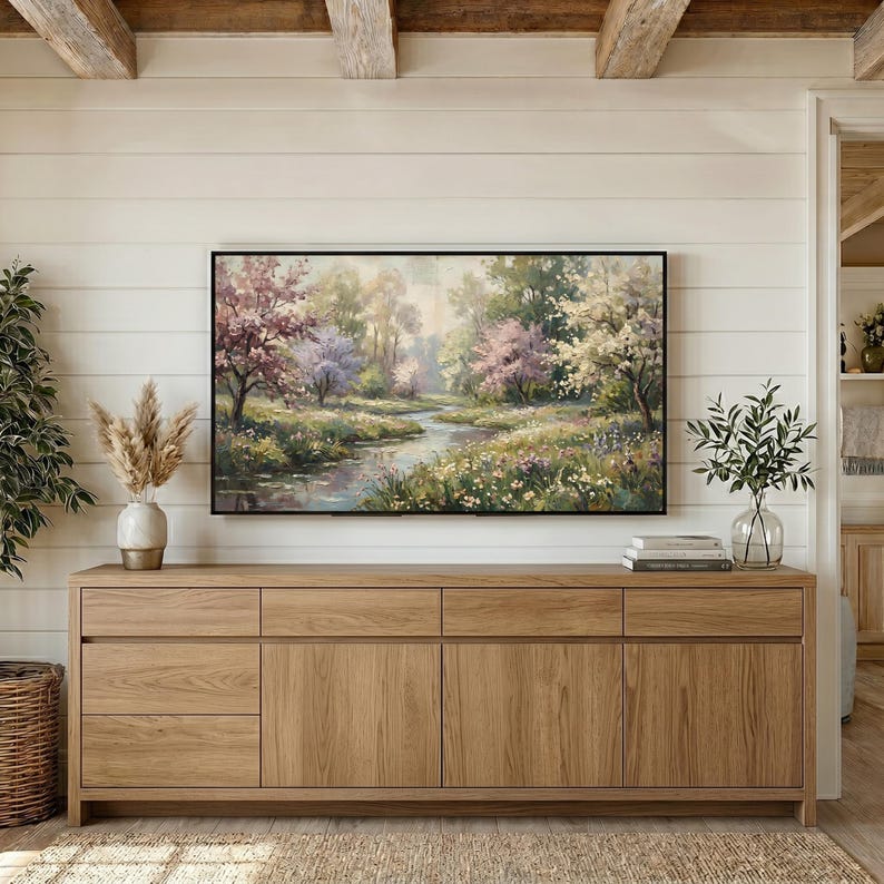 A cozy country farmhouse living room showcasing a tranquil spring meadow landscape painting on a digital TV display. The artwork is mounted on a bright white shiplap wall over a long natural oak wood media console table. A stylish woven basket with dried pampas grass and modern vases complete the warm, inviting setup. Redefine your home display now—click for an instant digital download.