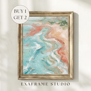 May include: Framed abstract art print depicting a beach scene with turquoise water, coral-colored sand, and small figures of people surfing. The artwork is in a gold-colored frame. The text "BUY 1 GET 2" is in a white circle in the upper left corner. The text "EXAFRAME STUDIO" is at the bottom.