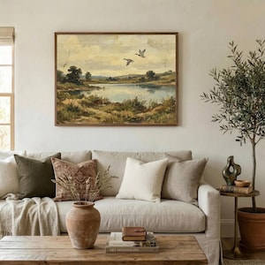 [Muted Country Rustic Farmhouse Wall Art] displayed in an organic modern living room above a beige linen sofa with earthy-toned pillows. The scene is bright and airy, featuring an olive tree in a terracotta pot and natural textures that complement the flying ducks landscape print. This printable nature art creates a warm, welcoming environment for grandmothers and new homeowners alike — instant digital decor for any mood.