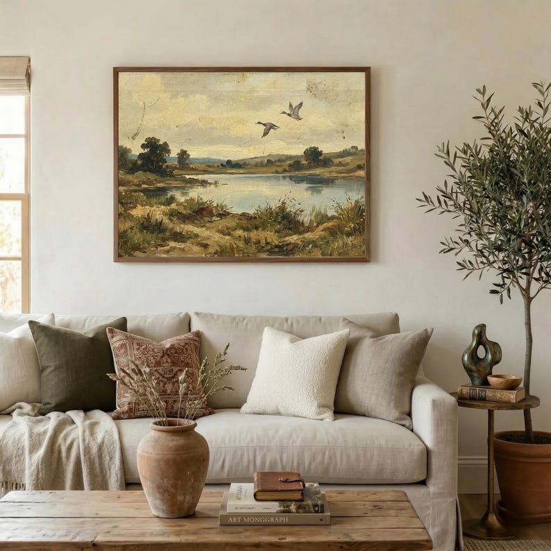 [Muted Country Rustic Farmhouse Wall Art] displayed in an organic modern living room above a beige linen sofa with earthy-toned pillows. The scene is bright and airy, featuring an olive tree in a terracotta pot and natural textures that complement the flying ducks landscape print. This printable nature art creates a warm, welcoming environment for grandmothers and new homeowners alike — instant digital decor for any mood.