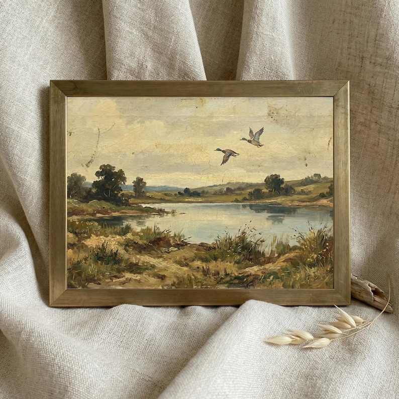 [Vintage Duck Art] presented in a thin minimalist gold frame resting on a soft, draped linen backdrop. The close-up view emphasizes the muted, earthy tones of the mallard ducks and the atmospheric depth of the wetland landscape. This elegant presentation highlights the "Fine Art" quality and museum-worthy finish of the digital download, making it a perfect gift for art collectors — download beautiful art for your home instantly.