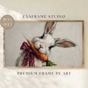 May include: A framed print of a white rabbit holding a carrot in its mouth, with a burgundy bow around its neck. The artwork has a neutral background and the text "EXAFRAME STUDIO" and "PREMIUM FRAME TV ART" are visible.