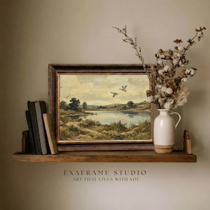 [Vintage Duck Art] elegantly positioned on a rustic dark wood floating shelf alongside antique books and a ceramic pitcher of dried cotton branches. The mallard ducks antique oil painting stands out with its warm earthy tones and heritage aesthetic against a neutral wall. This lifestyle mockup captures the essence of cozy country home decor, perfect for a study or library — download beautiful art for your home today.
