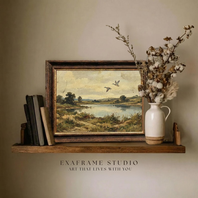[Vintage Duck Art] elegantly positioned on a rustic dark wood floating shelf alongside antique books and a ceramic pitcher of dried cotton branches. The mallard ducks antique oil painting stands out with its warm earthy tones and heritage aesthetic against a neutral wall. This lifestyle mockup captures the essence of cozy country home decor, perfect for a study or library — download beautiful art for your home today.