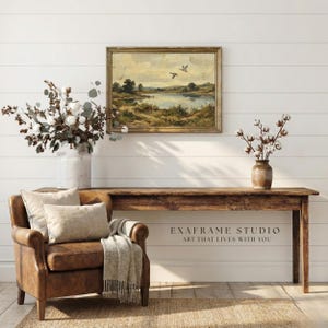 [Vintage Flying Ducks Landscape Print] displayed in an elegant wooden frame above a rustic timber console table and a classic brown leather armchair. The scene features a cozy living room with bright white shiplap walls, natural cotton branches, and a woven jute rug. This lifestyle mockup captures the restorative "Modern Heritage" vibe of the mallard ducks antique oil painting with its visible brushstrokes — bring calm artistry to your walls.