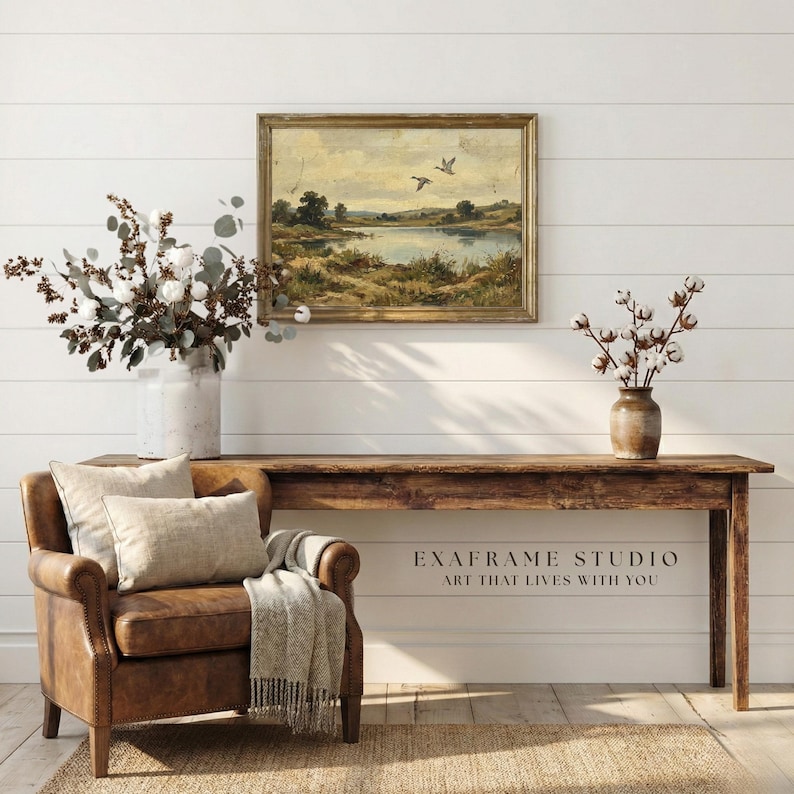 [Vintage Flying Ducks Landscape Print] displayed in an elegant wooden frame above a rustic timber console table and a classic brown leather armchair. The scene features a cozy living room with bright white shiplap walls, natural cotton branches, and a woven jute rug. This lifestyle mockup captures the restorative "Modern Heritage" vibe of the mallard ducks antique oil painting with its visible brushstrokes — bring calm artistry to your walls.