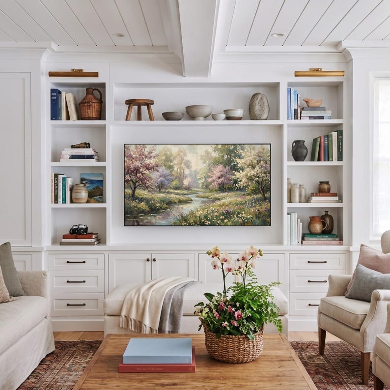 A luxurious custom living room featuring built-in white bookshelves framing a tranquil spring meadow TV art display. The cozy central seating area includes a wooden coffee table styled with books and a fresh floral basket, creating a sophisticated gallery wall ambiance. Transform your entertainment center into a fine art showcase—click to download your digital file now.