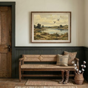 [Moody Lake House Decor] featured in a sophisticated entryway with dark green wood-paneled walls and a rustic woven wooden bench. The vintage flying ducks landscape print is mounted in a wide wooden frame, paired with leather riding boots and a basket of cotton branches. This moody nature print creates a serene, grounded atmosphere in any entryway or lodge — bring calm artistry to your walls with this instant digital download today.