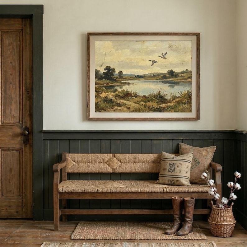 [Moody Lake House Decor] featured in a sophisticated entryway with dark green wood-paneled walls and a rustic woven wooden bench. The vintage flying ducks landscape print is mounted in a wide wooden frame, paired with leather riding boots and a basket of cotton branches. This moody nature print creates a serene, grounded atmosphere in any entryway or lodge — bring calm artistry to your walls with this instant digital download today.