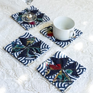 May include: Four square fabric coasters with a pinwheel design. Each coaster has a navy blue and white striped border with a central patchwork design of various colours. A wine glass and a white mug rest on two of the coasters.