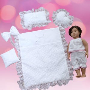 May include: A doll wearing a white textured sleeveless top and matching shorts with pink trim, accompanied by a white bedding set including a rectangular pillow, a heart-shaped pillow, a small square pillow, a large blanket, and a bow.