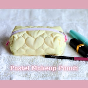 May include: A pale yellow quilted makeup pouch featuring a heart-shaped pattern and a light purple zip closure. The text "Pastel Makeup Pouch" is printed at the bottom. A black eyeliner pencil and two lipstick tubes are positioned nearby.