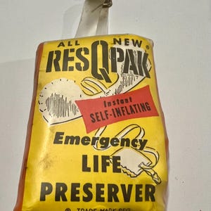 May include: A vintage RES-Q-PAK emergency life preserver. The yellow and red packaging features the text "ALL NEW RES-Q-PAK" and "Instant Self-Inflating". A metal clip and white strap are attached to the top of the package.