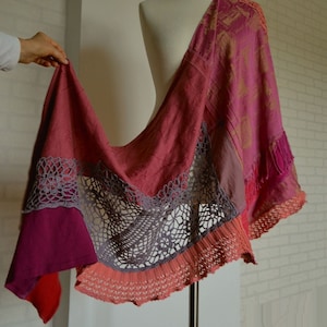 Puede incluir: Un chal drapeado en tonos de rosa, rojo y morado, exhibido en un maniquí. El chal presenta un diseño de patchwork con detalles de encaje de ganchillo en gris y coral. Los bordes están acabados con flecos.