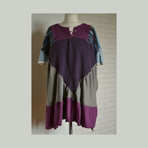 Könnte beinhalten: Ein Patchwork-Kleid mit einzigartigem Design. Das Kleid kombiniert lila, graue und blaue Stoffe. Das Oberteil hat eine strukturierte lila Strickpartie, die Ärmel sind blau-lila gestreift. Das Kleid wird auf einer Büste präsentiert.