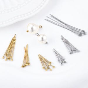May include: A collection of jewellery making supplies including two white faux pearl charms with metal loops, several bundles of gold-toned ball-end headpins, and multiple bundles of silver-toned ball-end headpins arranged on a white surface.