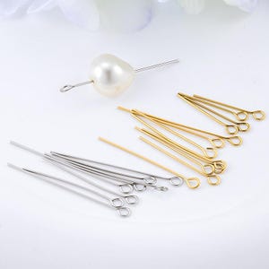 May include: A collection of silver and gold-toned metal eye pins for jewellery making, featuring thin straight shafts with a small circular loop at one end. One silver eye pin is threaded through a single, smooth, off-white, oval-shaped pearl bead.