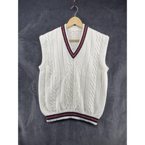 May include: A cream-colored sleeveless sweater vest featuring a V-neckline and a cable knit pattern. The neckline and hem are accented with a striped trim in dark red and navy blue. The garment is displayed on a wooden hanger against a dark background.